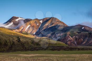  In the hollows of rhyolitic mountain snow. Above the source of hot water steam rises. Early summer morning in the National Park Landmannalaugar, Iceland