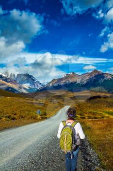 Woman tourist with a backpack is on the roadside. The gravel road goes into the distance. Mountains and rocks in Torres del Paine National Park, Chile. The concept of active and extreme tourism