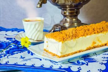 Professional bakery. Gorgeous white cheesecake, sprinkled with sweet crumbs. The background is a shiny samovar and an porcelain cup with hot tea on blue kitchen towel