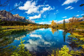 Concept hiking. Bright shining day in the Canadian Rockies. Canmore, near Banff. Jagged mountains and red-orange trees are reflected in smooth water of the lake