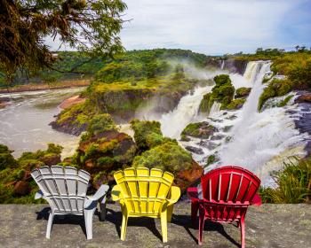 Three plastic armchairs - chaise longue - white, yellow, red - set at waterfalls. Scenic basaltic rock formations famous waterfalls Iguazu Falls. The concept of ecological and phototourism