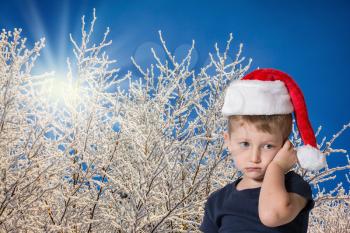 The child has sad blue eyes and fair soft hair. The charming little boy in red cap of Santa Claus. Photo executed on background of snow-covered forest