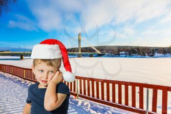 The bridge and the frozen river. Concept of active winter tourism. Adorable little boy with blue eyes in a red Santa Claus hat