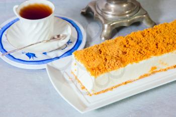 Professional bakery. Exquisite white cheesecake, sprinkled with sweet orange crumbs. The background is porcelain white cup with hot tea and samovar on blue kitchen towel