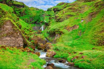 Basalt mountains covered in green grass and moss. Gorgeous cascading waterfall from melting glacier. Iceland, July