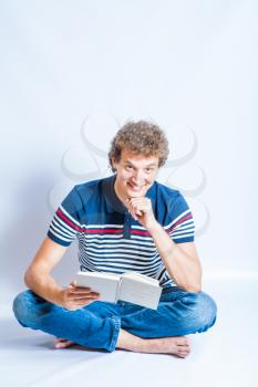 Smiling young man sitting on a floor and reading a book. Modern student with curly hair. Studio shot on the gray background, vertical composition