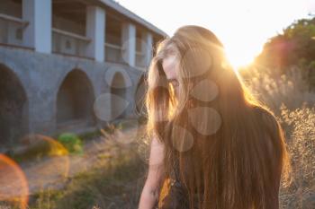 Blonde girl at in sunrise light. Face half hidden by long hair. Citypark with ancient building on background. Backlit with lens flare