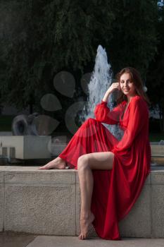 Young women in red vintage dress sitting on fountain parapet and looking at camera.