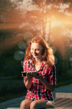 Redhead girl sitting in city park and having fun communicate via tablet PC backlit by sunset