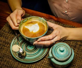 Lady holding cup of milk coffee in cafe over the sourcer with metal spoon