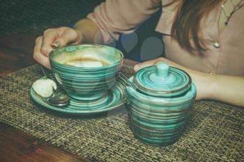 Lonely lady drinking coffee in the morning, side view of female hands holding stripped cup of hot beverage on wooden desk. There is a sugar bowl aside.