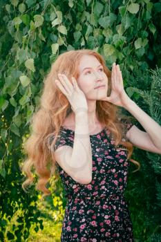 Portrait of a serious beautiful redhead lady in summer dress fixing her hair with hands under birch tree in spring time.
