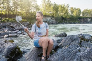Woman taking selfie on mobile phone with stick on mountain river background