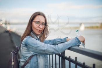 Young beautiful woman with bottle of the water staying on embankment in blurred background