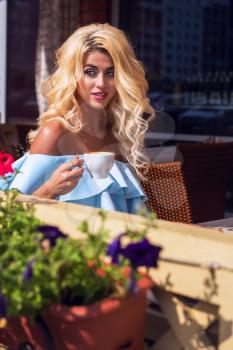 Beauty sensual woman in elegant blue dress in cafe. Young woman sitting in cafe with cup of coffee in summer day.