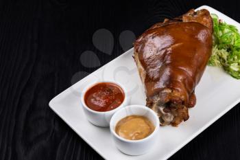 Tasty pork knuckle with sauces and vegetables on a black wooden background