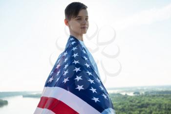 Blonde boy waving national USA flag outdoors over blue sky at the river bank. Beauty summer sunny day. American flag, patriotism, independence day 4th july concept