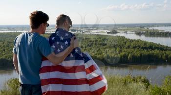 Father with American flag embraces his son on river bank. Patriotism, independence day 4th july concept