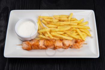 Grilled salmon shashlik with fried potatoes on white plate on black wooden background