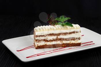 Plate with piece of delicious caramel cream cake decorated with mint leaves on black wooden background