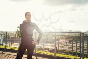 A woman in sportswear with bottle of water have rest at sunset on the evening city background