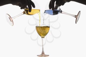Two hands pouring different colored cocktails into a wine glass