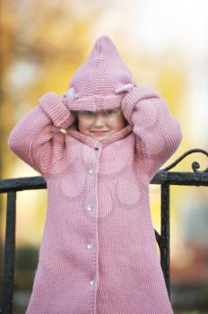 A little blonde girl 3-4 years old in a knitted coat with a hood stands on a bench against the background of an autumn Park during a walk