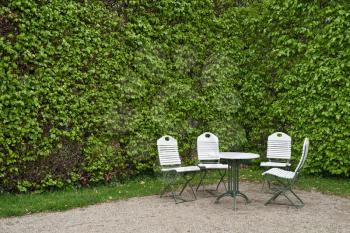 White garden furniture chairs and a round table against a hedge wall with text space in a European public park