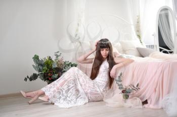 Photo of a young girl in the style of fayn art. A girl in a white translucent dress sits on the floor next to the bed and a bouquet of flowers.