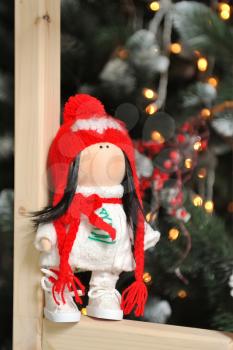 A cute children's soft doll in a red hat stands next to a Christmas tree. New Year's gift children's soft doll