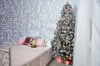 Interior against the background of a white wall with cold garlands, a forged bed and a Christmas tree. Garland on the whole wall, place for text