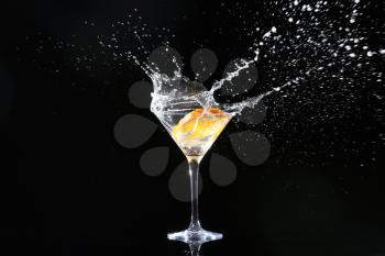 Glass with splashing cocktail and slice of orange on black background�