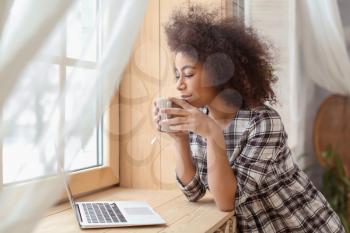 Beautiful African-American woman drinking tea while using laptop at home�