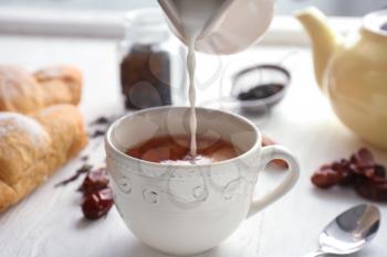 Pouring milk into cup with aromatic tea on table�