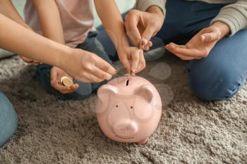 Little girl with her parents sitting on carpet and putting coin into piggy bank indoors. Money savings concept�