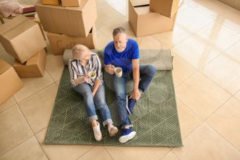 Mature couple with belongings sitting on carpet and drinking tea indoors. Moving into new house�