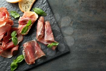 Slate board with tasty prosciutto slices and basil on table�