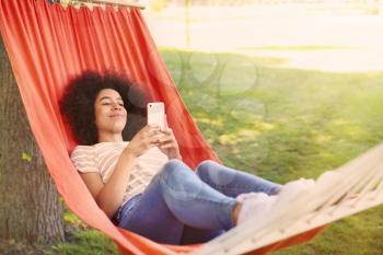Beautiful young African-American woman with mobile phone resting in hammock outdoors�