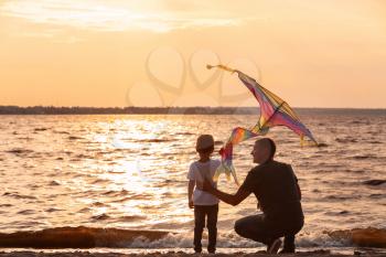 Happy father and son flying kite near river at sunset�