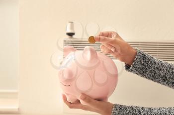 Woman putting coin into piggy bank near radiator at home. Heating saving concept�