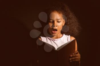 Surprised African-American girl reading book on dark background�