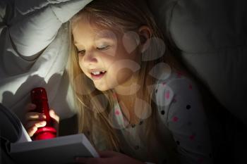 Cute little girl with flashlight reading book in bed under blanket�