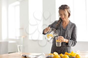 Young woman pouring fresh lemonade from jug into glass at home�