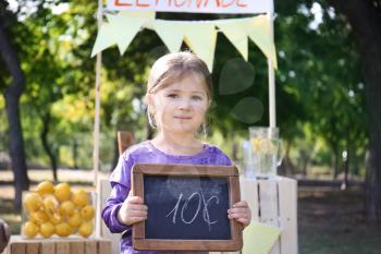 Little girl holding small blackboard with price near lemonade stand in park�
