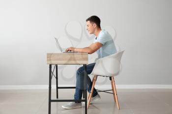 Young man working with laptop while sitting at table against light wall�