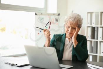 Senior woman suffering from headache while working with laptop indoors�