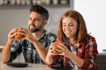Young couple eating tasty burgers at home�