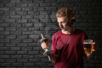 Young man with dumbbell and mug of beer near black brick wall. Concept of choice between alcohol and sport�