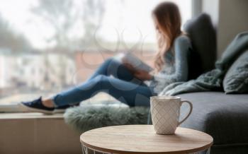 Cup on table of young woman reading book on window sill�