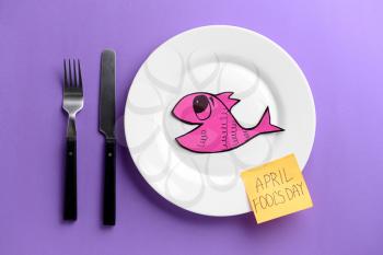 Sheet of paper with words April fools food, fish, plate and cutlery on color background�